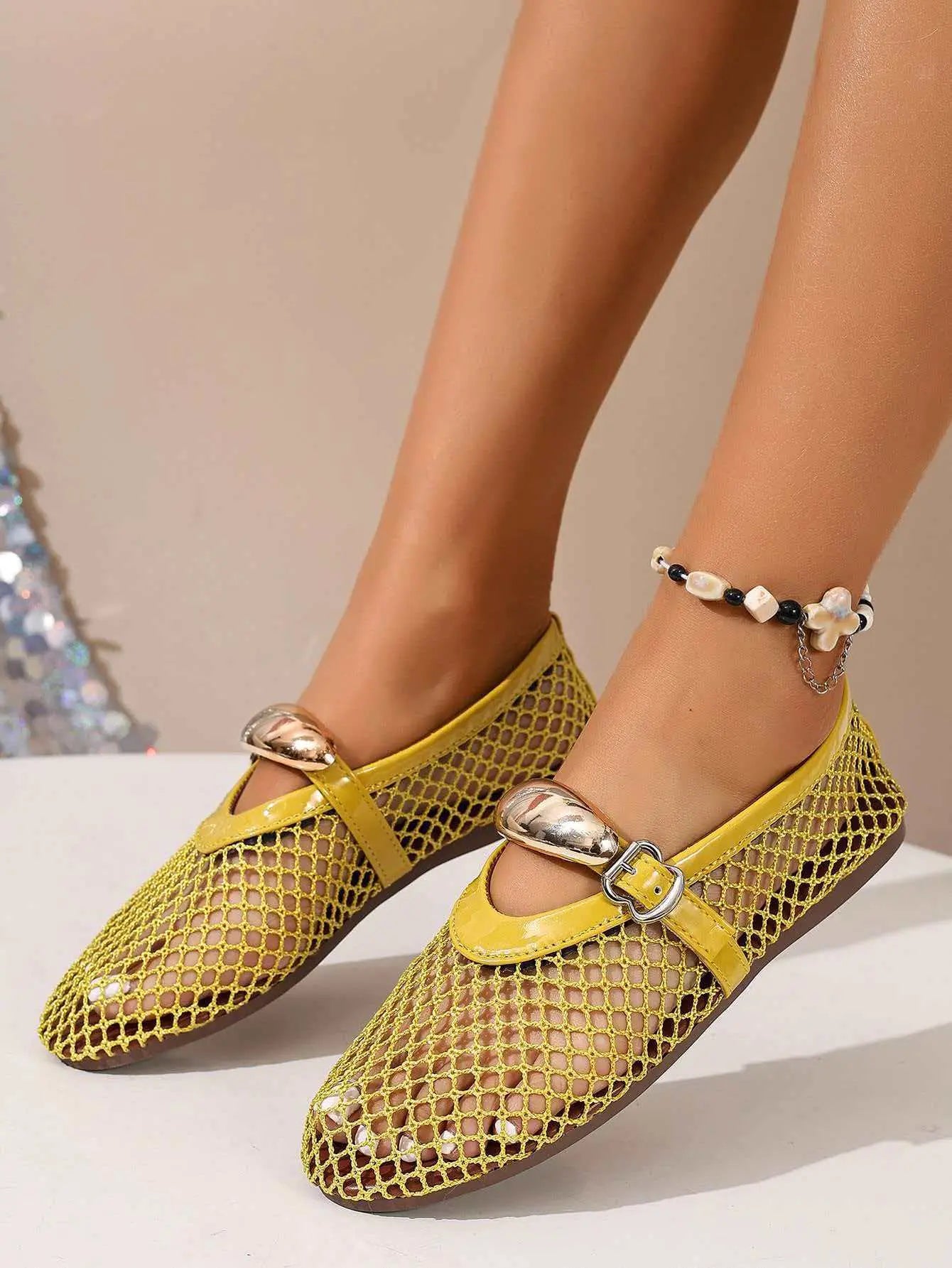 A pair of bright yellow mesh flat sandals with patent faux leather trim and decorative metallic oval buckles, shown on a woman's feet with a beaded anklet. The airy design is perfect for summer.