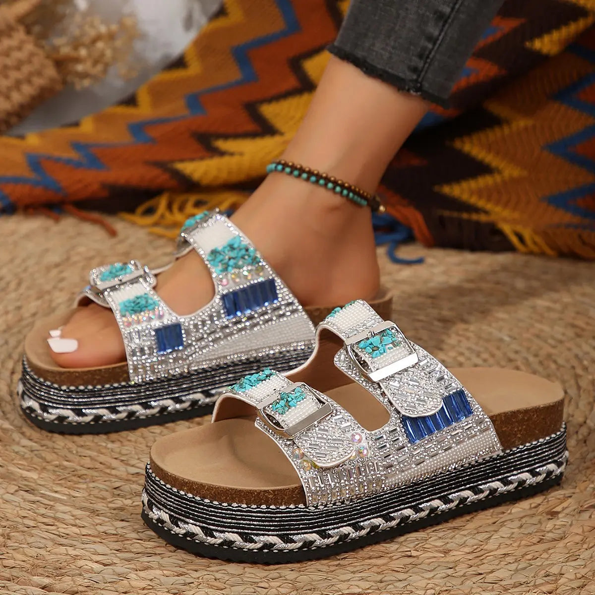 Sorami embellished platform slide sandals with double buckle straps featuring rhinestones, blue, and turquoise stones, worn by a model with a bohemian-inspired outfit and anklet.