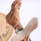 Women's beige wedge sandal with a single clear PVC strap featuring a gold pearl accent, worn on a model's foot with a sparkling anklet against a white background.