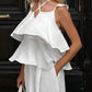 A woman models an elegant white linen set featuring a tiered ruffle spaghetti strap top and flowing wide-leg pants, styled with a straw bag in an outdoor urban setting.