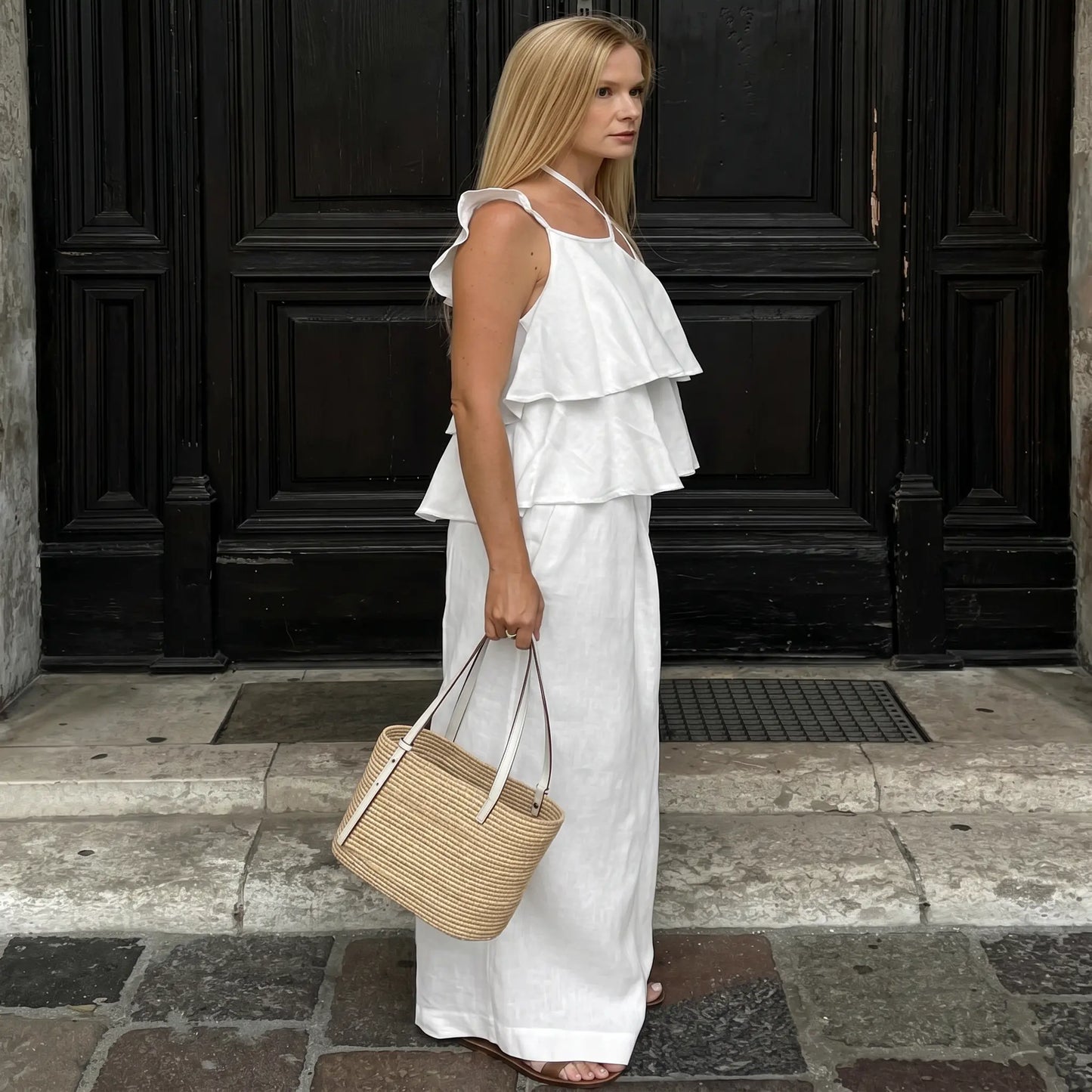 Woman modeling a flowing white linen set featuring a tiered ruffle spaghetti strap top and wide-leg pants, shown from the side in an outdoor setting.