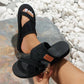 Black Sorvan women's casual chic platform slide sandals with criss-cross fabric straps and a distressed 'FASHION' label, shown on a foot with white toenails against a natural stone and pebble background.