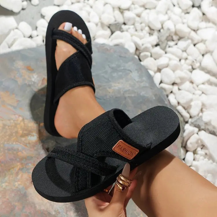Black Sorvan women's casual chic platform slide sandals with criss-cross fabric straps and a distressed 'FASHION' label, shown on a foot with white toenails against a natural stone and pebble background.