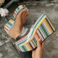 Close-up of a woman's feet wearing vibrant, multi-colored striped platform wedge sandals with a clear PVC strap, set against a textured outdoor ground.