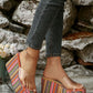 Model wearing vibrant multi-colored striped platform wedge slide sandals with a clear PVC strap, paired with dark wash jeans, set against a rustic stone wall.