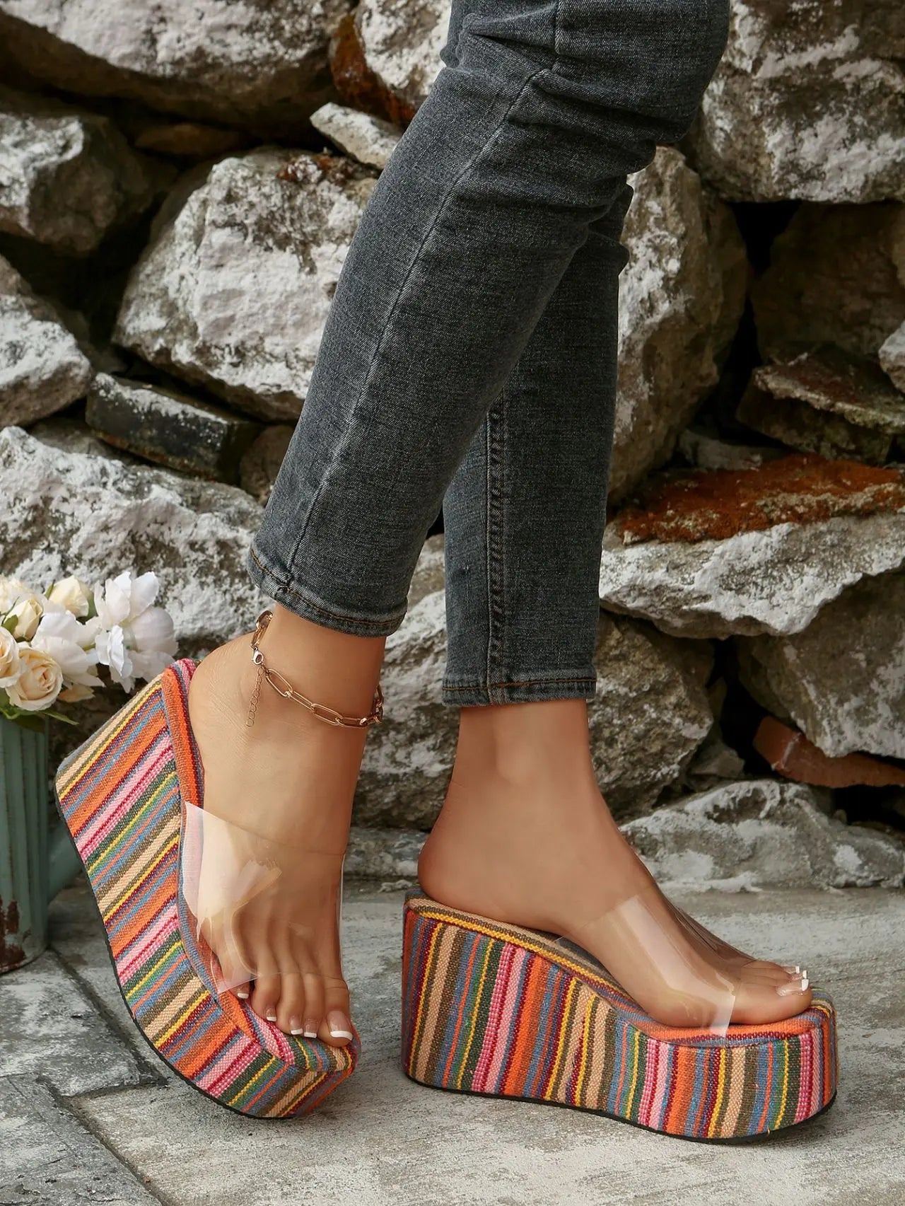 Model wearing vibrant multi-colored striped platform wedge slide sandals with a clear PVC strap, paired with dark wash jeans, set against a rustic stone wall.