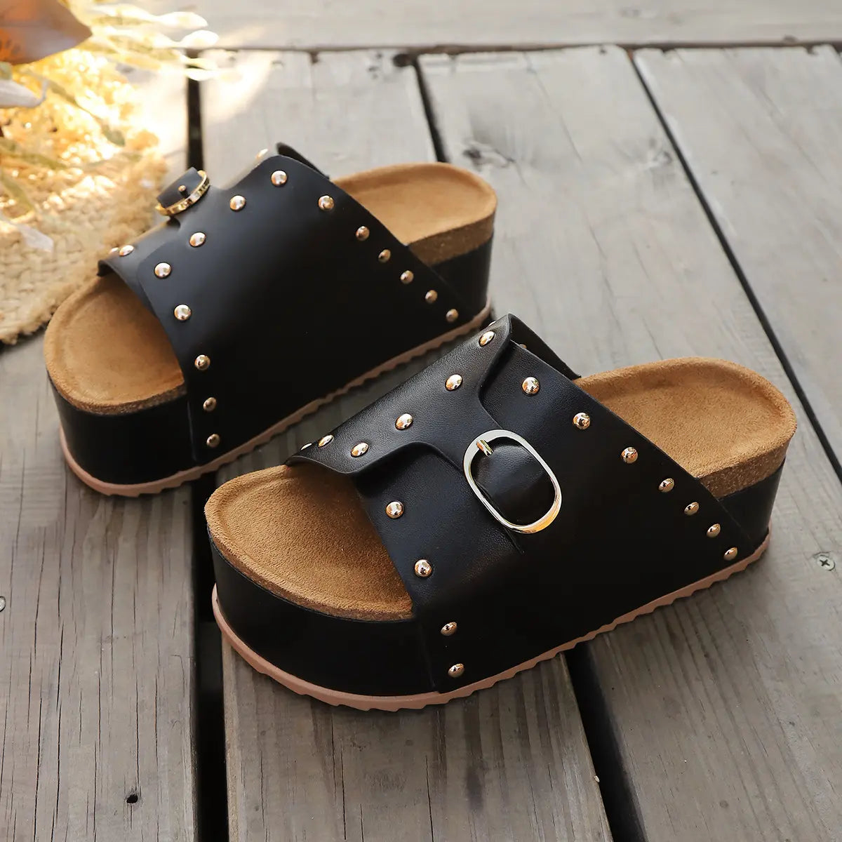 Black platform slide sandals with gold stud embellishments and a cork-effect insole, shown from a three-quarter angle on a wooden surface.