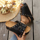 Black studded platform slide sandals with a cork-effect insole and gold buckle, shown on feet with a straw hat in the background.