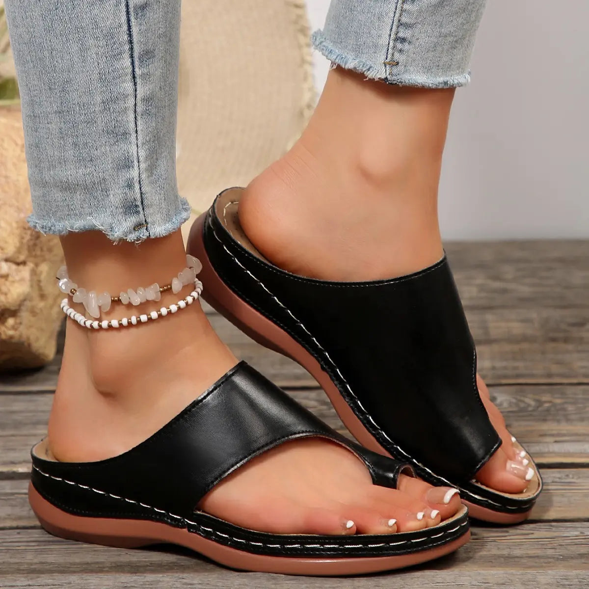 Black PU leather casual slip-on slides with toe-post design worn by a woman wearing ripped jeans and a beaded anklet on a wooden floor.