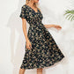 A woman models a V-neck midi dress in the 'Midnight Bloom' ditsy floral print, featuring short flutter sleeves and a cinched smocked waist, set against a textured white background with plant shadow.