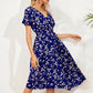 A woman wears a navy blue midi dress with a ditsy floral print, featuring a V-neck, short flutter sleeves, and a cinched smocked waist, shown in a three-quarter view against a white studio background with subtle shadow play.