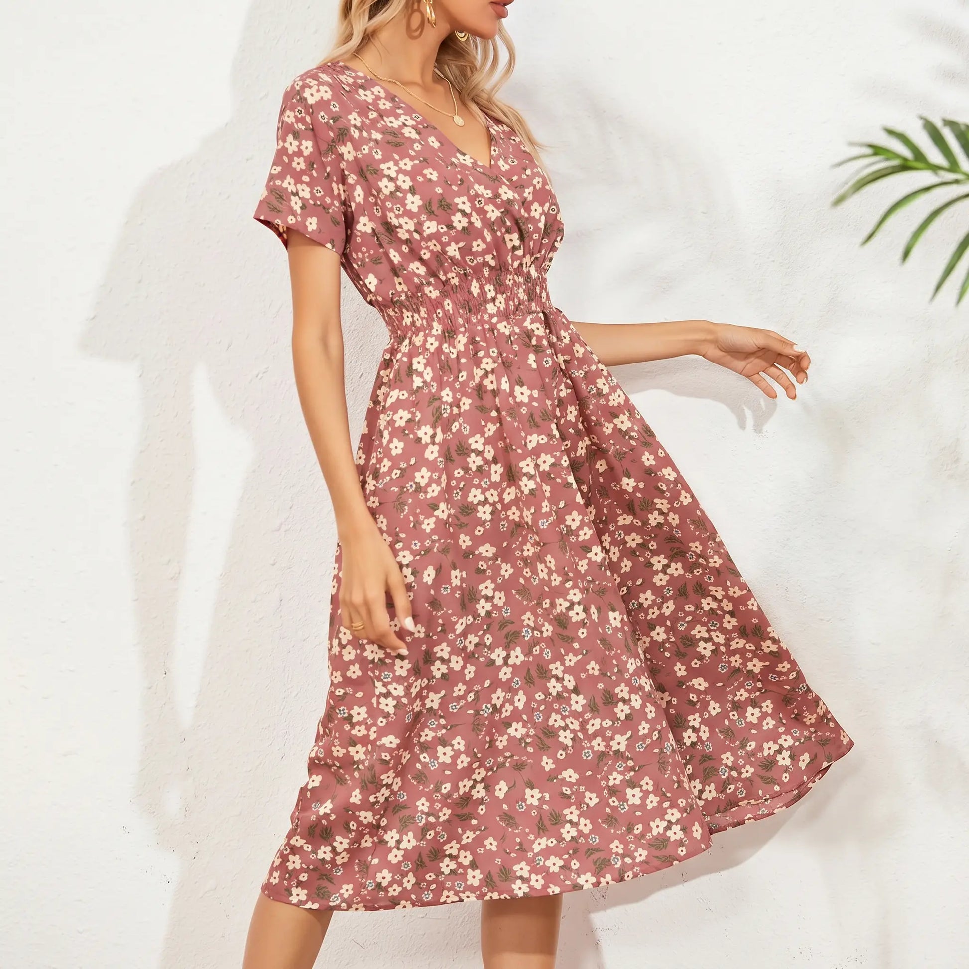 A woman wears a rose blush midi dress with a ditsy floral print, featuring a V-neck, short flutter sleeves, and a smocked elastic waistband, styled for a bohemian-romantic look. Full view of the A-line skirt.