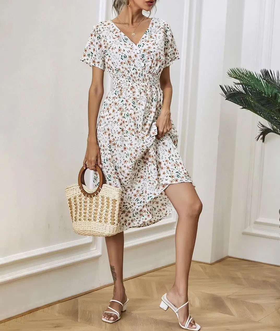 A woman models the Talore white V-neck midi dress featuring a ditsy floral print, short flutter sleeves, and a cinched waist, accessorized with a straw bag and white sandals. This lifestyle shot showcases the dress's flowy silhouette in a natural setting.