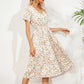 A woman models a white midi dress featuring a ditsy floral print, V-neck, and flutter sleeves, shown against a textured wall with palm leaf shadows.
