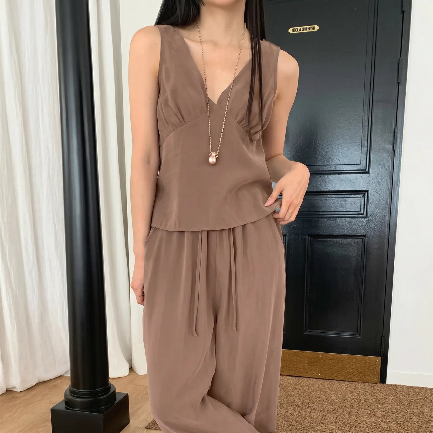 A woman models a muted taupe sleeveless V-neck crepe set featuring a gathered bust top and flowy wide-leg drawstring trousers, styled with a long necklace.