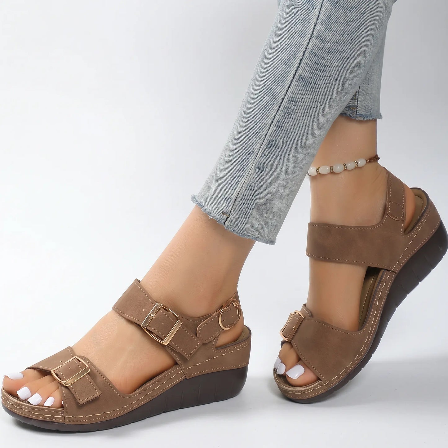 Fashionable taupe faux suede wedge sandals with gold buckles and white contrast stitching, worn by a woman in jeans on a studio background. Features adjustable straps for comfort and style.