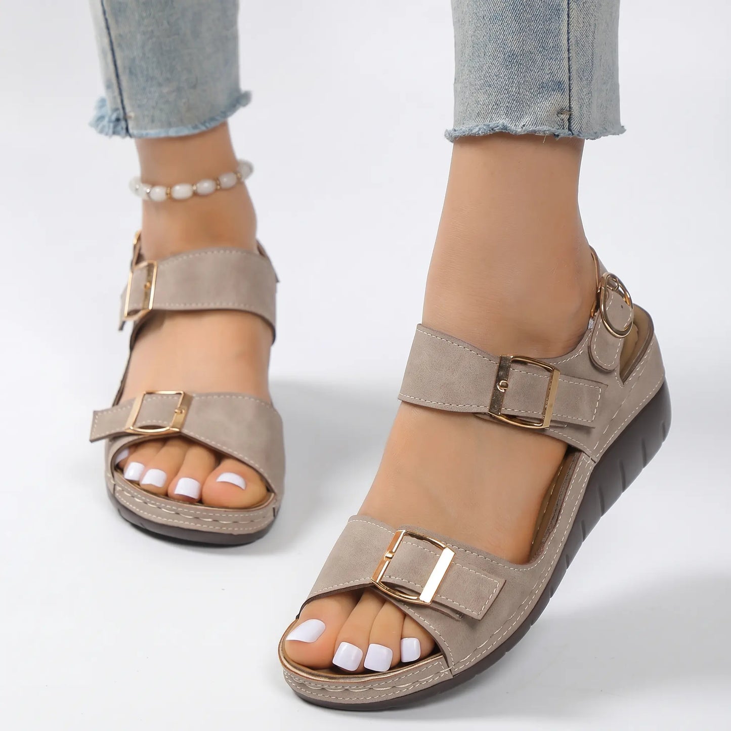 Comfortable taupe faux suede wedge sandals for women, featuring dual forefoot straps with gold buckles and an adjustable ankle strap, worn on feet with light wash jeans.