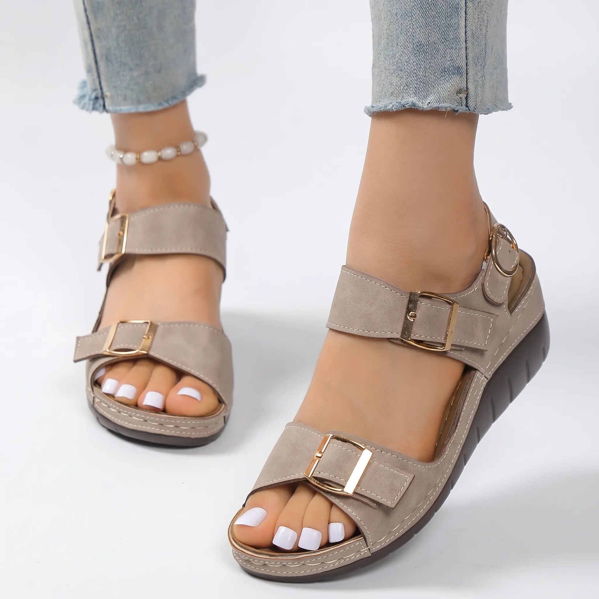 Comfortable taupe faux suede wedge sandals for women, featuring dual forefoot straps with gold buckles and an adjustable ankle strap, worn on feet with light wash jeans.