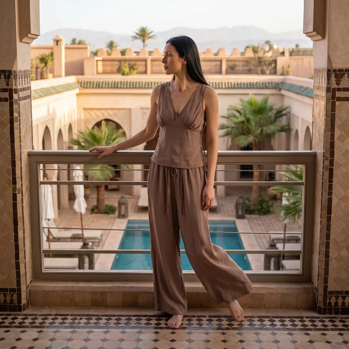 A woman stands on a balcony overlooking a Moroccan riad courtyard, wearing a flowing taupe V-neck sleeveless top and wide-leg trousers.