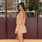A woman models a taupe halter neck mini dress with a smocked waist and flowy skirt, seen from a three-quarter side view outdoors.