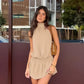 A woman wears a flowing taupe halter neck mini dress with a smocked waist, styled for a sophisticated bohemian summer look.