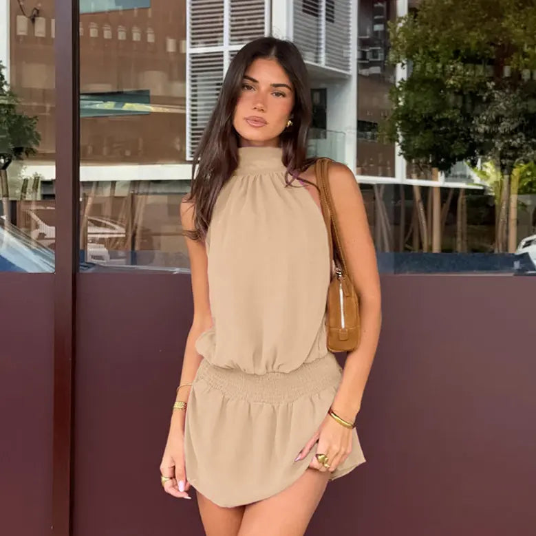 A woman wears a flowing taupe halter neck mini dress with a smocked waist, styled for a sophisticated bohemian summer look.