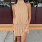 A woman models a taupe halter neck mini dress with a smocked waist, styled casually outdoors with a shoulder bag. This bohemian dress drapes fluidly for a romantic look.