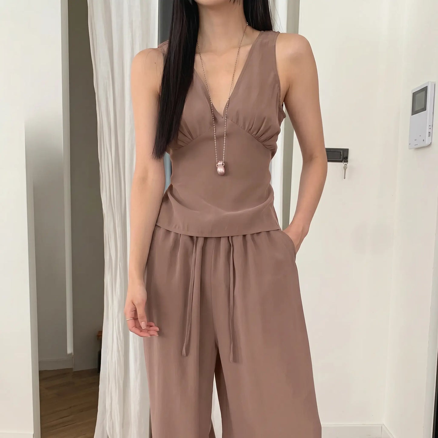 A woman models a taupe two-piece set featuring a sleeveless V-neck top with gathered bust detail and flowy wide-leg trousers with a drawstring waist.