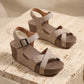 Taupe faux leather wedge sandals with criss-cross straps, gold-toned studs, and a speckled brown platform sole, displayed on a textured surface.