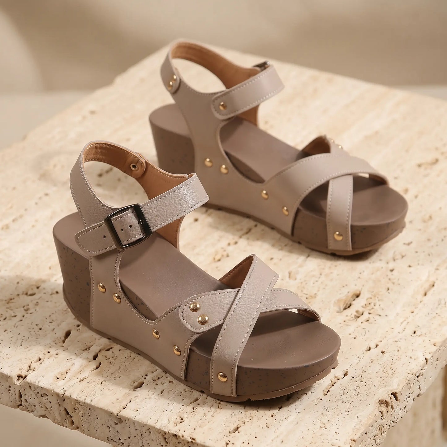 Taupe faux leather wedge sandals with criss-cross straps, gold-toned studs, and a speckled brown platform sole, displayed on a textured surface.