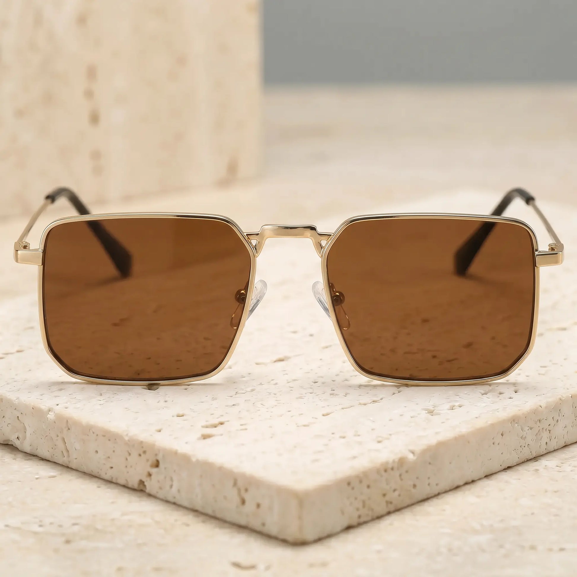 Front three-quarter view of Tavren vintage square sunglasses with gilded gold metal frames and mocha brown tinted lenses, resting on a textured stone surface.