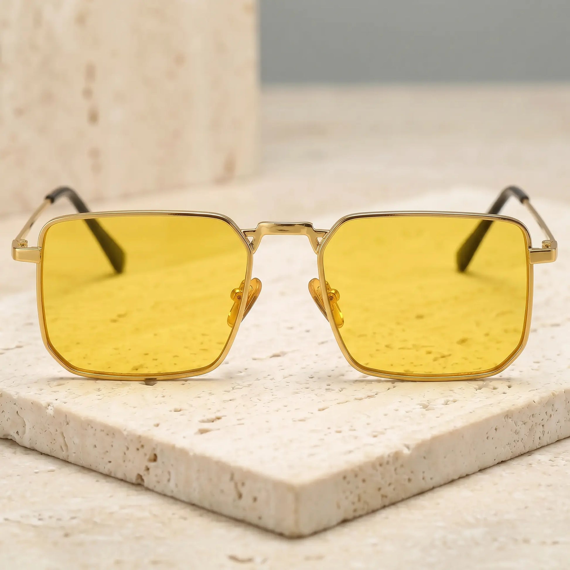 Tavren gilded gold square sunglasses with amber yellow lenses. Features thin metal frames, clear nose pads, and black temple tips, displayed on a textured stone.