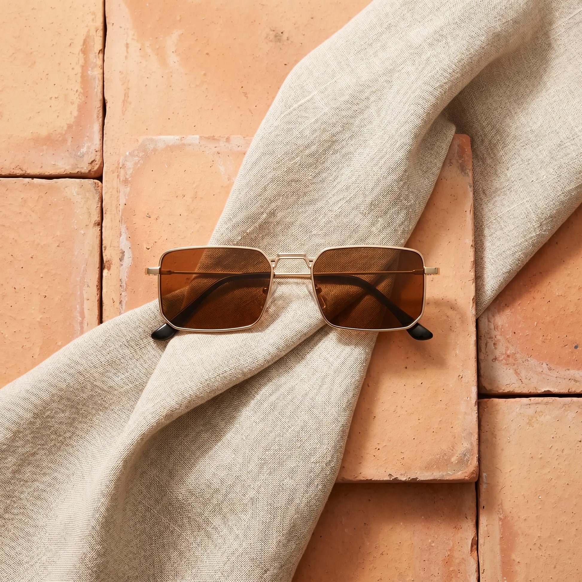 Tavren vintage square sunglasses with gilded gold ultra-thin metal frames and rich brown lenses, artfully arranged on a light beige linen cloth over rustic terracotta tiles.