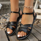 Black braided flat sandals with an adjustable ankle strap, worn by a model with white nail polish, styled with a gold chain anklet on a wooden deck with pebbles.