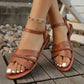 Lifestyle image of Tavri warm brown flat braided summer sandals worn by a model, featuring an open-toe design with braided and smooth faux leather straps and an ankle strap, set against a rustic wooden deck and stone background.