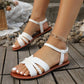 White flat bohemian sandals with braided straps and an adjustable ankle strap, worn by a model in a natural outdoor setting on wooden planks and pebbles.