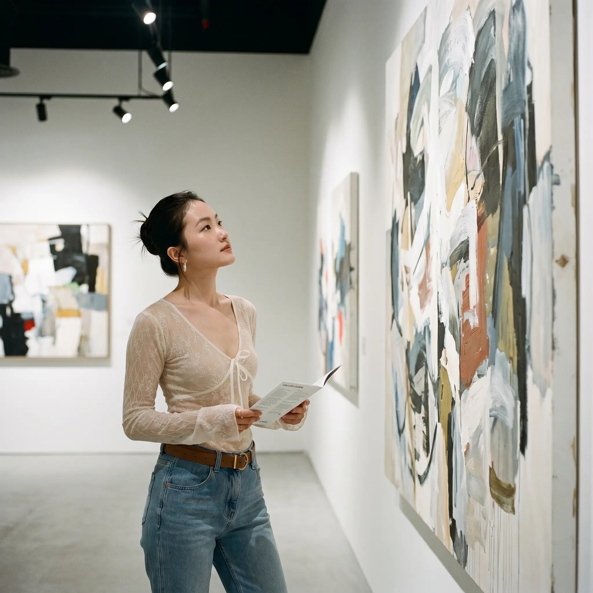 Woman wears a Tazuri sheer vintage cream floral lace tie-front top with a deep V-neck and flared sleeves, styled with blue jeans, admiring art in a gallery.