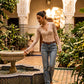 Model wears a Tazuri sheer floral lace tie-front top in vintage cream with blue jeans, touching a fountain in a sunlit Moroccan Riad.