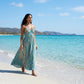 Woman walking barefoot on a sunny beach, wearing a flowing teal halter maxi dress with an artisanal tie-dye print. The gathered neckline and open back design are visible as she strolls beside turquoise ocean waters.