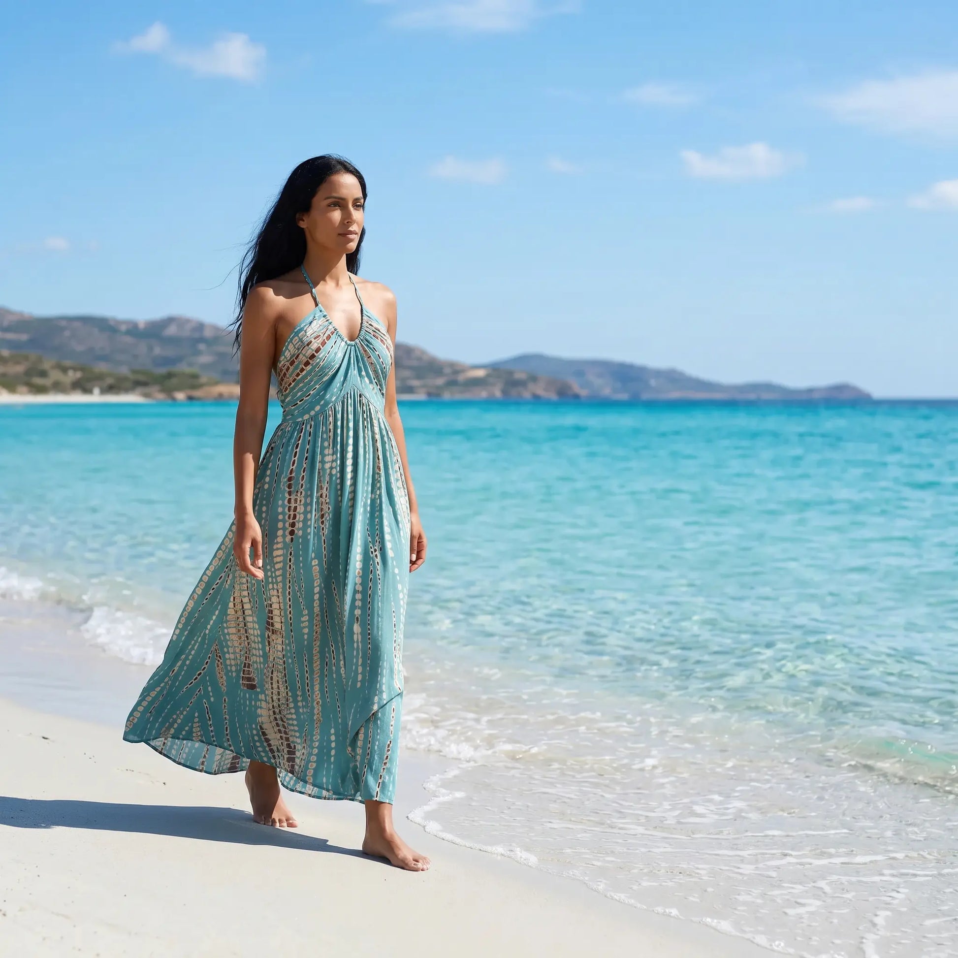 Woman walking barefoot on a sunny beach, wearing a flowing teal halter maxi dress with an artisanal tie-dye print. The gathered neckline and open back design are visible as she strolls beside turquoise ocean waters.