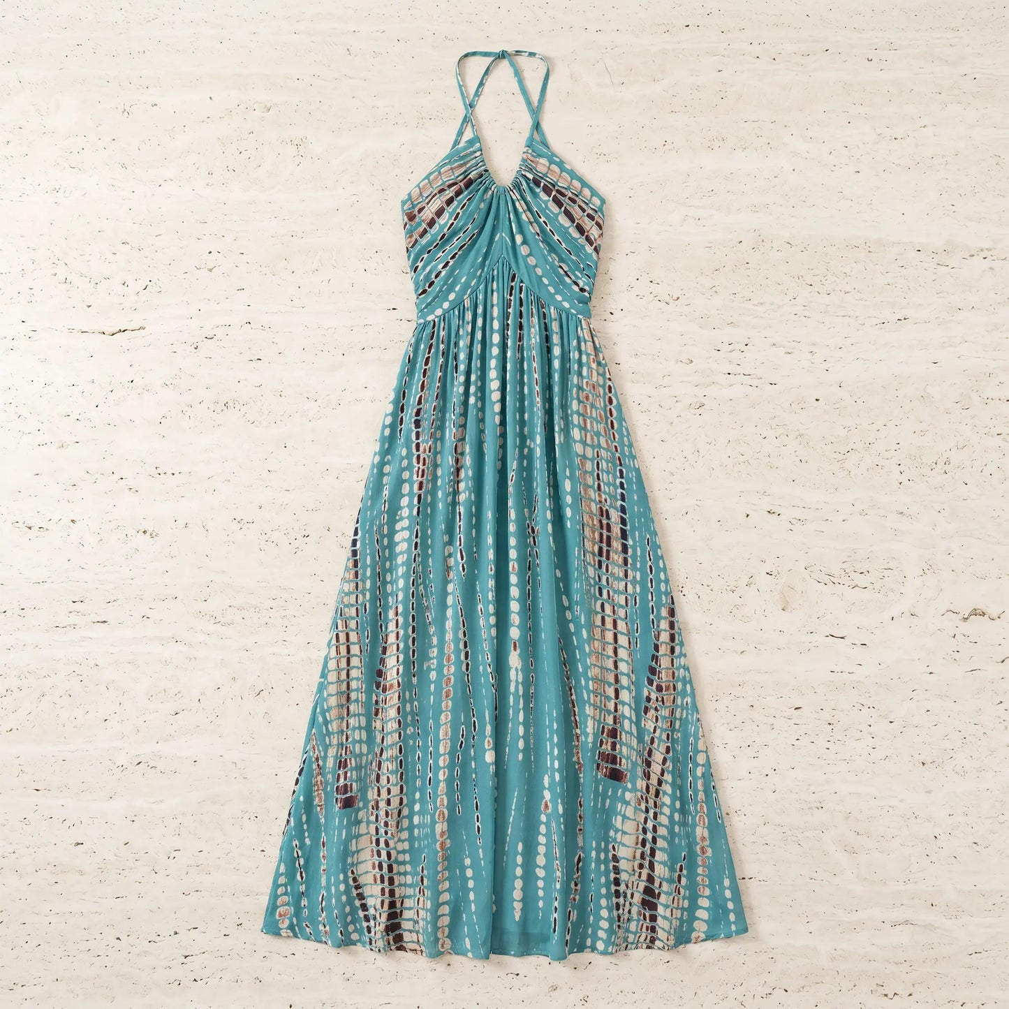 Teal tie-dye halter maxi dress, laid flat. Features a gathered neckline, flowing A-line silhouette, and vertical pattern of white ovals and brown dotted stripes.