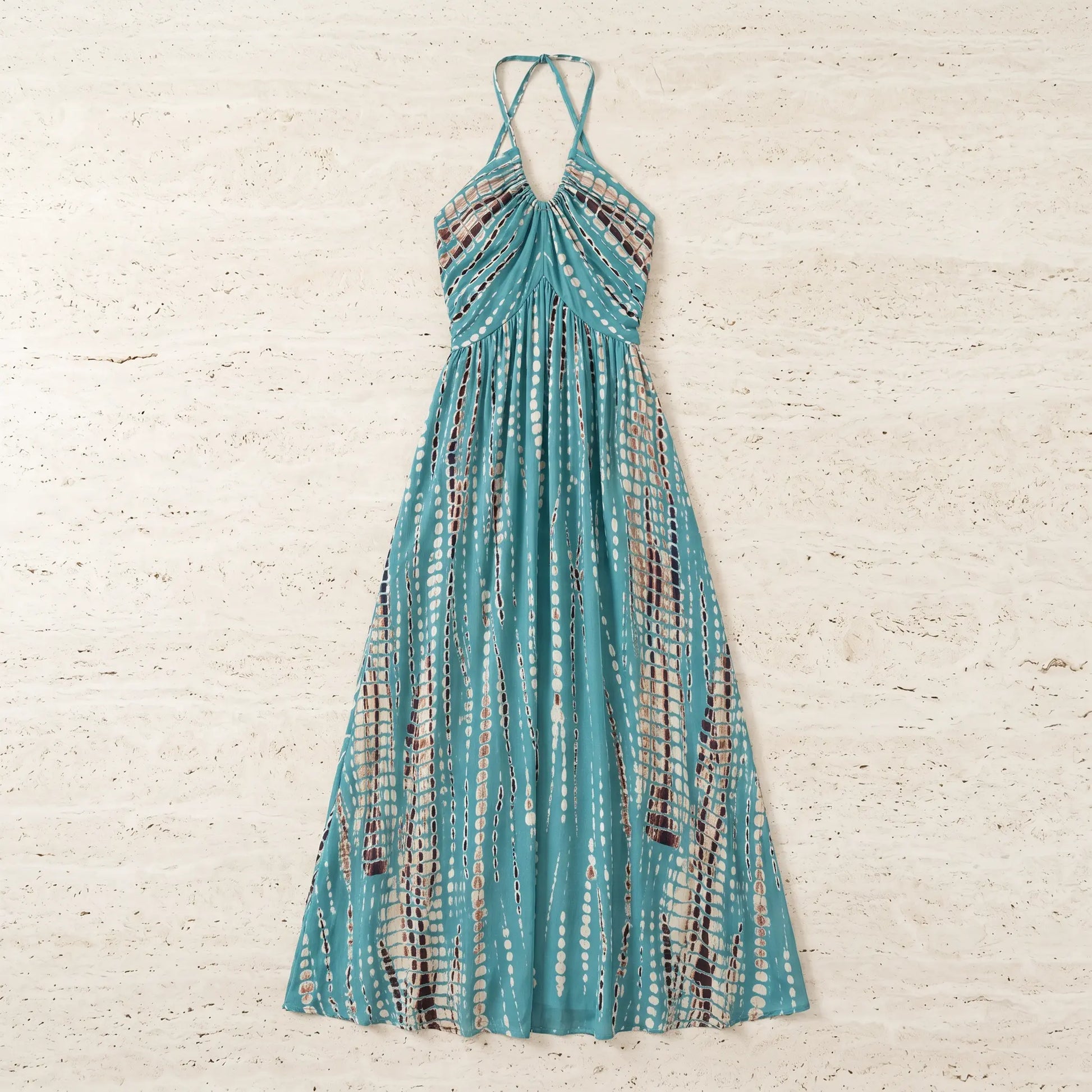 Teal tie-dye halter maxi dress, laid flat. Features a gathered neckline, flowing A-line silhouette, and vertical pattern of white ovals and brown dotted stripes.
