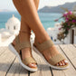 A woman wears Tenzun Sand Khaki woven elastic platform sandals, featuring wide, textured straps and a thick white platform sole. Her feet are crossed on a wooden deck, overlooking a picturesque blue sea and white village buildings.