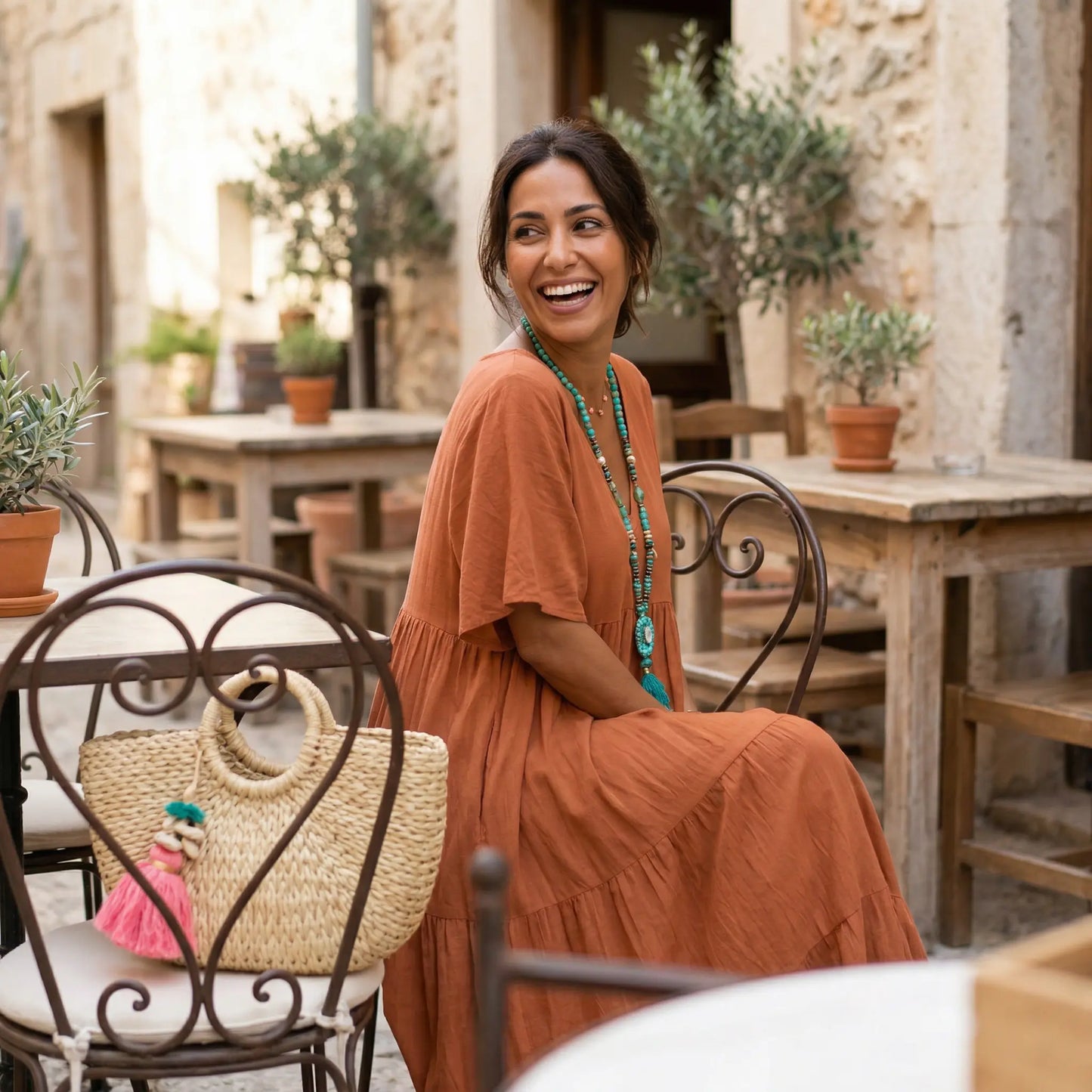 Smiling woman wearing a terracotta crinkled gauze tiered maxi dress with short sleeves and a V-neck, sitting at an outdoor cafe. She wears a turquoise necklace.