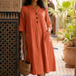 Woman models a vibrant terracotta V-neck midi dress with crinkled fabric, black button placket, half sleeves, and large front patch pockets, standing in a beautiful tiled courtyard.