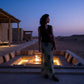 Female model in Thalwyn dark navy V-neck knit top and sage green abstract mesh maxi skirt, captured at dusk by an outdoor fire pit.