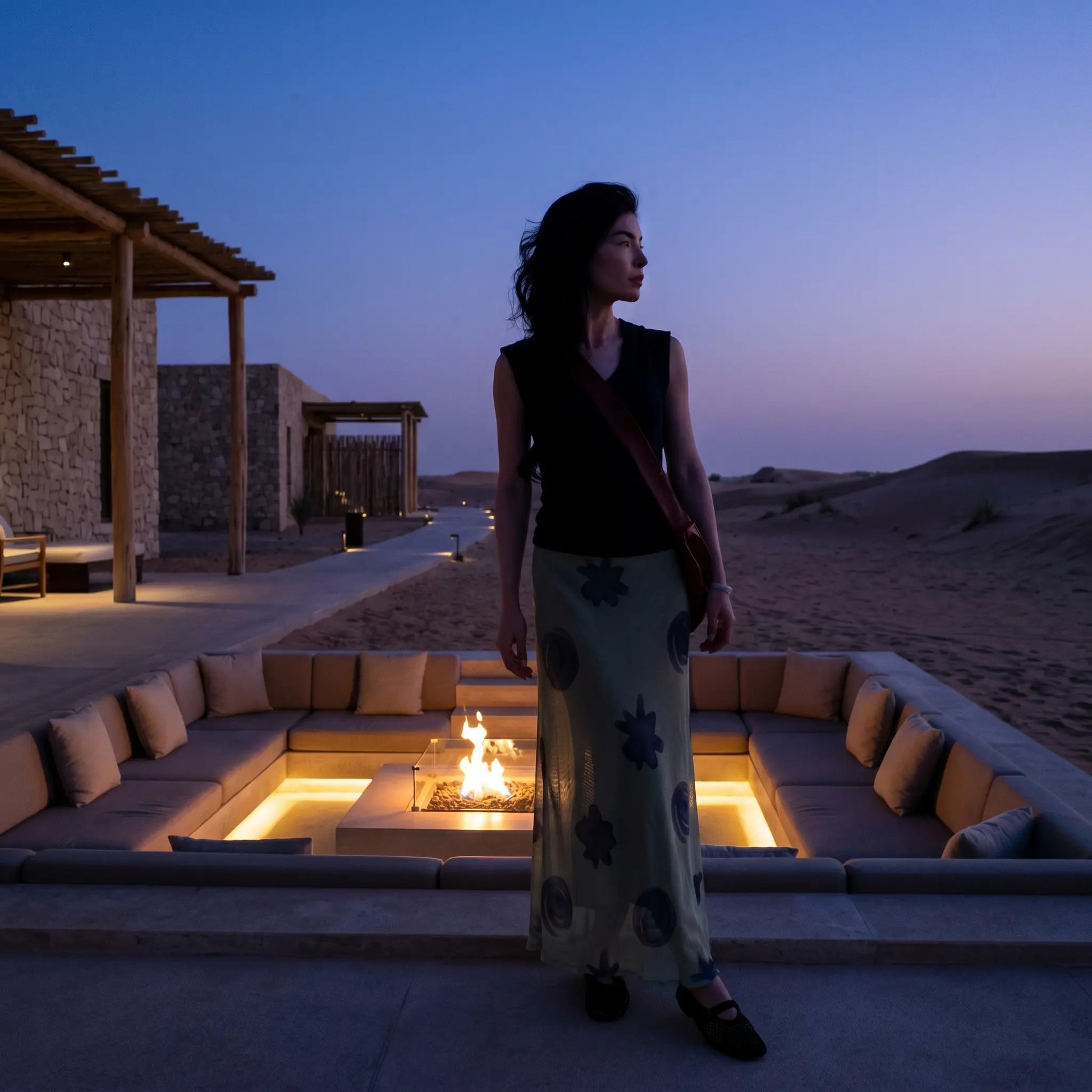 Female model in Thalwyn dark navy V-neck knit top and sage green abstract mesh maxi skirt, captured at dusk by an outdoor fire pit.