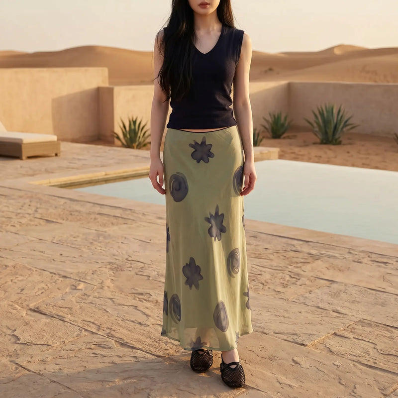 Female model in Thalwyn navy V-neck knit top and sage green abstract mesh maxi skirt, showcasing the flowing silhouette and unique patterns in a warm desert resort setting.