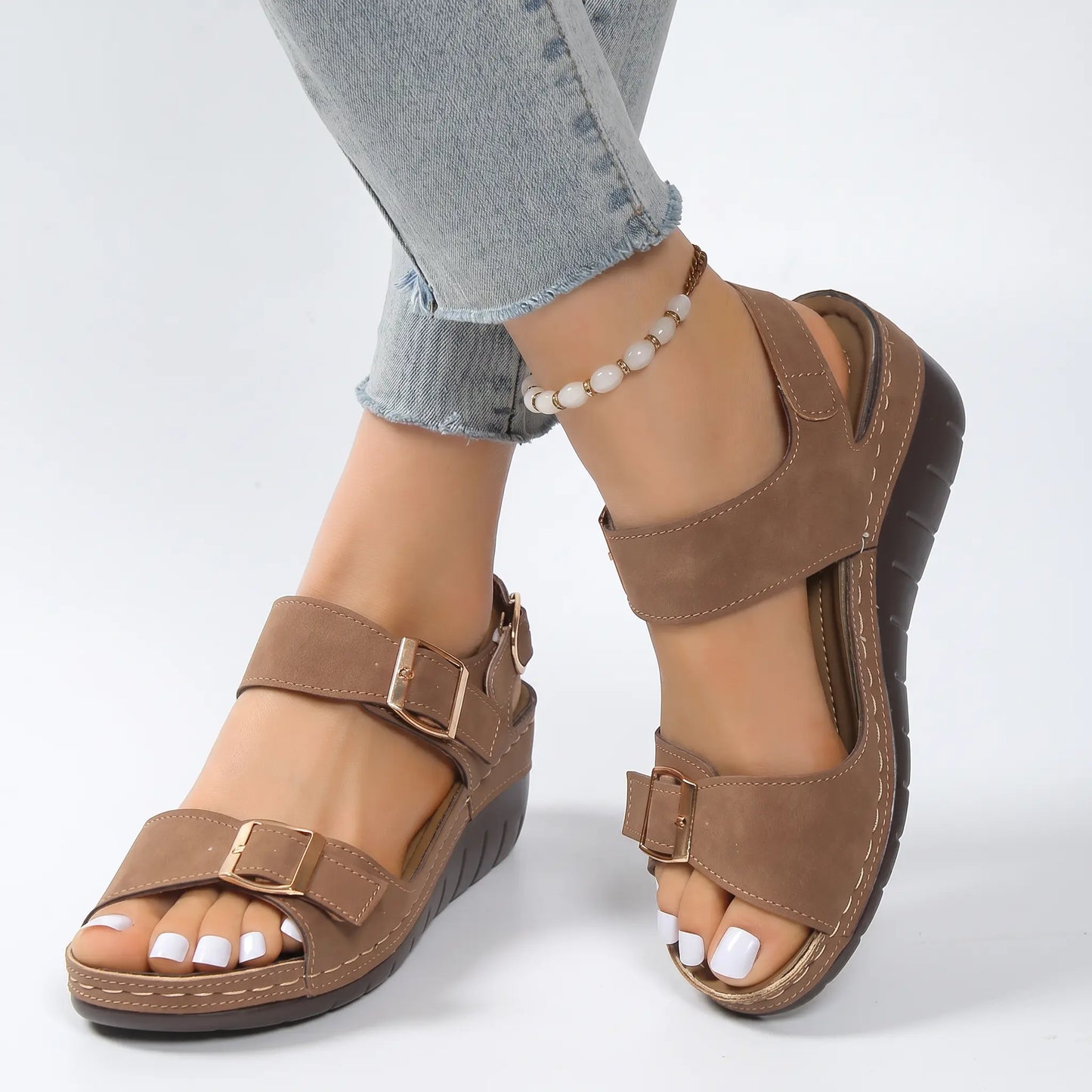 Torvin women's wedge sandals in a versatile tan faux suede, featuring two adjustable forefoot straps with gold buckles and an ankle strap, paired with denim jeans. These comfortable strappy sandals showcase white contrast stitching and a textured wedge heel.