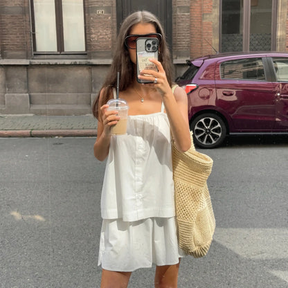Woman wearing a white 100% cotton camisole and relaxed shorts set outdoors, holding a drink and a straw bag. The camisole features spaghetti straps and a square neckline with button detailing.